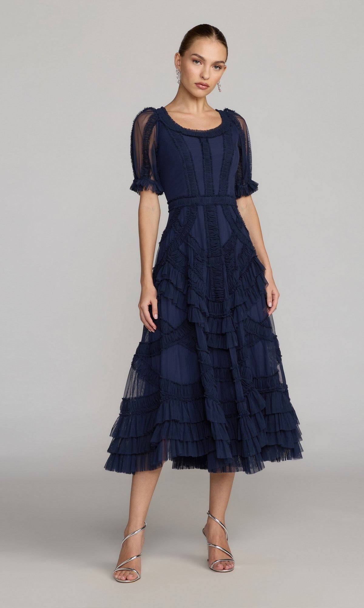 A woman in the Indigo Mac Duggal 8248 Midi Wedding Guest Dress with ruffles, sheer sleeves, and silver heels stands against a plain background—an elegant choice for wedding guests.